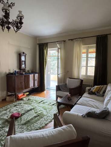 A cozy living room with white couches, wooden furniture, green patterned rug, vintage clock, and chandelier. Large windows with curtains let in natural daylight.