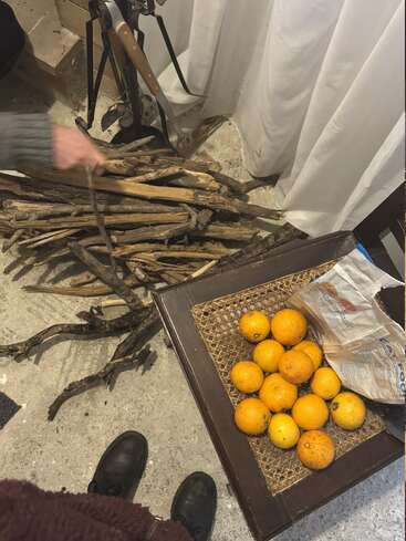A person arranges firewood beside white curtains. Oranges and an empty carton rest on a wooden chair. Concrete floor, black boots, and fireplace tools visible. Cozy atmosphere.