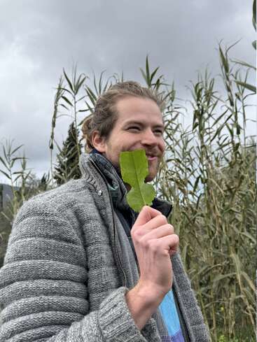 A man with a beard stands outdoors in front of tall grass, holding a green leaf to his face, smiling. He wears a grey sweater and looks playful.