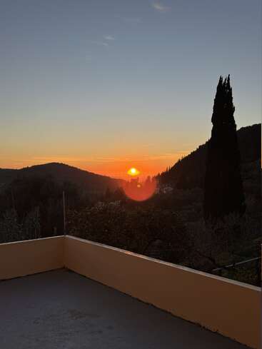 A stunning sunset over distant hills, orange hues filling the sky. Silhouetted trees and a simple terrace create a peaceful, serene early evening scene.