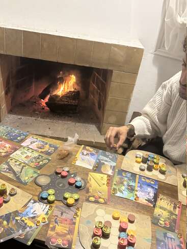 A cozy scene: a person in a white sweater plays a colorful board game beside a warm fireplace, surrounded by cards, tokens, and wooden game pieces.