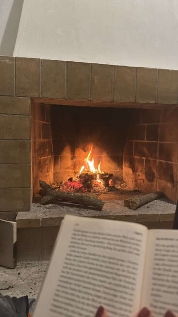 A cozy fireplace with a warm, crackling fire burns inside, while someone sits close, enjoying a book, creating a peaceful and relaxing atmosphere indoors.