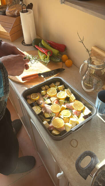 A person prepares a meal in a sunlit kitchen, adding rosemary to a baking tray with chicken, vegetables, and lemon slices beside fresh produce. Cozy atmosphere.