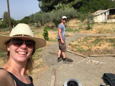 L'image montre une femme coiffée d'un chapeau de paille prenant un selfie avec un homme à l'arrière-plan, sur un chemin de terre dans un environnement rural.