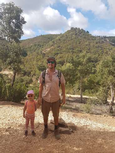L'image représente un homme et un enfant debout sur un chemin de gravier, sur fond d'arbres et de collines sous un ciel bleu avec des nuages blancs.