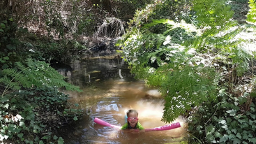 Une jeune fille nage dans une étendue d'eau, probablement un lac ou une rivière, avec une grande formation rocheuse en arrière-plan. L'eau est calme et paisible.