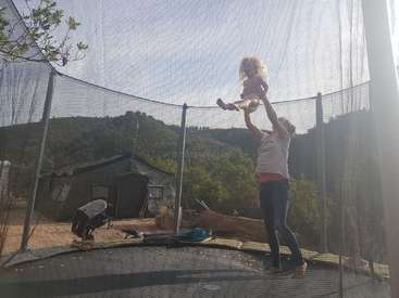 Une femme tient un enfant en l'air sur un trampoline, avec une maison et des arbres en arrière-plan par une journée ensoleillée.