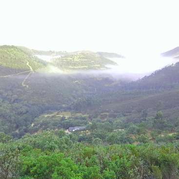 L'image représente un paysage montagneux serein avec une verdure luxuriante, une vallée brumeuse et un bâtiment lointain, le tout dans un ciel clair et couvert.