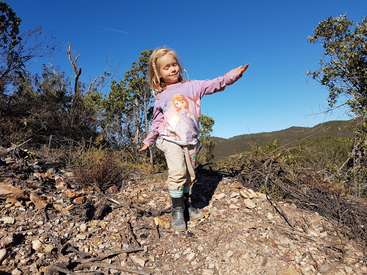 Une jeune fille aux cheveux blonds se tient sur une colline rocheuse, vêtue d'une chemise rose et de bottes, les bras tendus et un ciel bleu serein au-dessus d'elle.