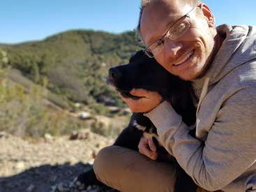 L'image représente un homme assis sur le sol, berçant un chien noir avec des marques blanches sur ses genoux, sur fond de colline pittoresque.