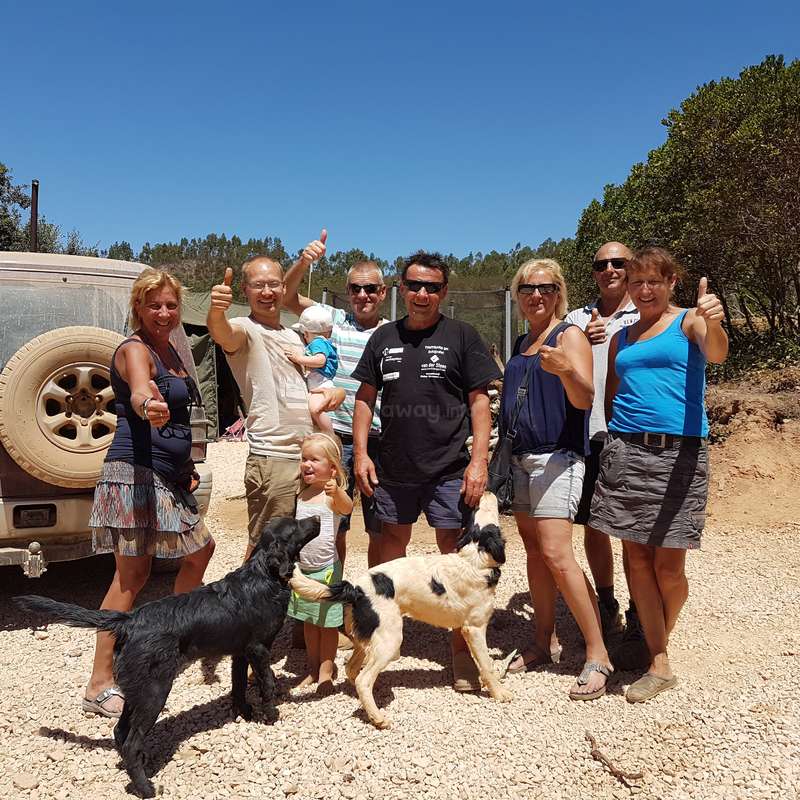 L'image montre un groupe de personnes et deux chiens posant pour une photo devant un véhicule, avec un ciel bleu clair et des arbres en arrière-plan.