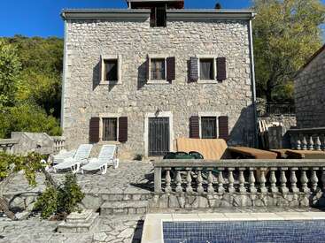 This image shows a rustic stone house with shuttered windows, a patio area with lounge chairs, balustrade fencing, and a swimming pool in the foreground.