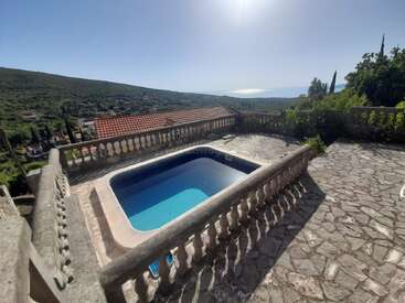 This image shows a stone terrace with a small empty pool, surrounded by a balustrade, overlooking green hills, rooftops, and a distant view of the sea.