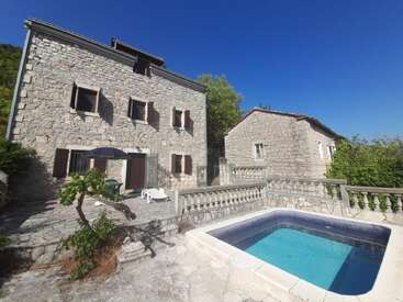 A charming stone house with wooden shutters, a spacious patio, sun loungers, umbrella, and a small square pool. Surrounded by nature under a clear blue sky.