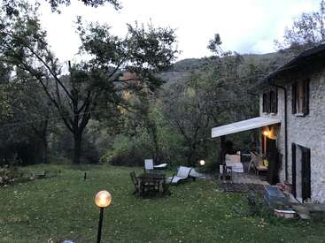 Une maison rustique en pierre avec un porche couvert se trouve au milieu des arbres et de la verdure. Des meubles d'extérieur et des lampes lumineuses créent une atmosphère de jardin paisible et accueillante à la tombée de la nuit.