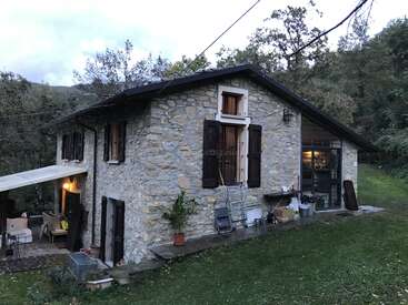 Une maison rustique en pierre avec des volets en bois se trouve au milieu d'une végétation luxuriante. Il y a un patio couvert avec des meubles d'extérieur, et des objets épars sont adossés au bâtiment.