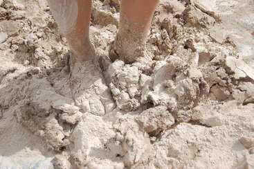 Un par de pies descalzos se hunden en barro espeso, húmedo y de color claro. El barro se pega a la piel, creando una escena al aire libre juguetona y desordenada.