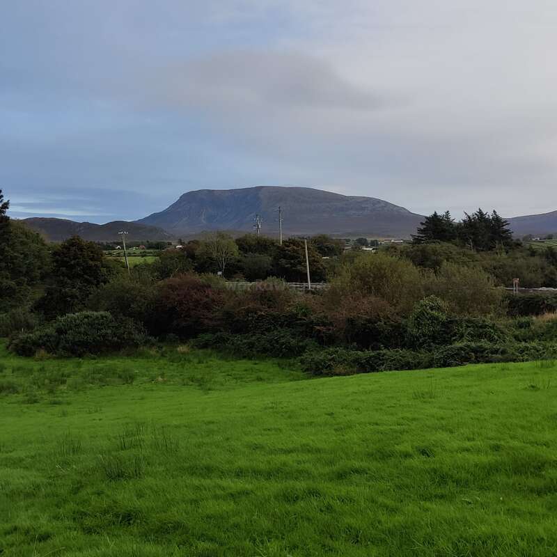 The image depicts a serene landscape featuring a lush green field, trees, and a distant mountain under a cloudy sky, evoking a sense of natural beauty and tranquility.