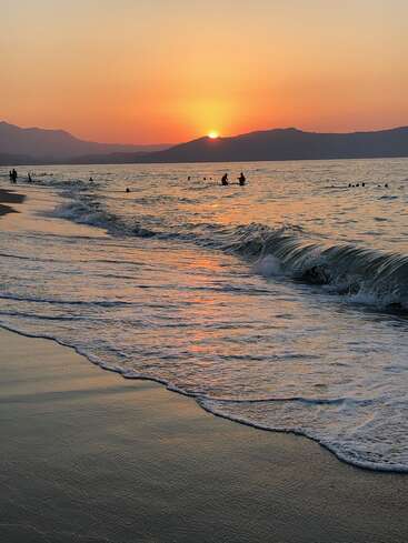 A imagem retrata uma cena serena de praia ao pôr do sol, com ondas suaves rolando na praia e pessoas nadando na água calma, em um cenário montanhoso.