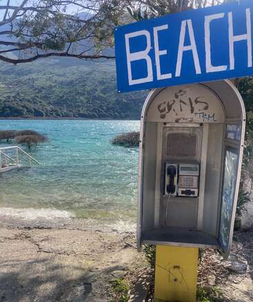 Uma cabine telefônica de praia com uma placa azul onde se lê \"BEACH\" fica na margem de um corpo de água, cercada por árvores e montanhas ao fundo.