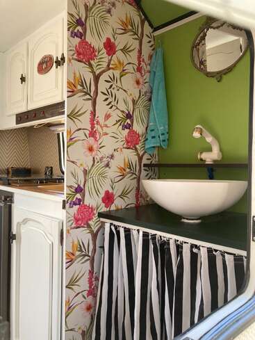 A imagem retrata o interior de uma van de camping, com uma pia, um espelho e um papel de parede floral, com uma cozinha e armários ao fundo.
