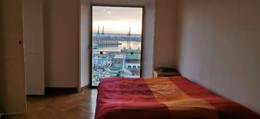 The image depicts a room with a large window, showcasing a cityscape outside. The interior features a table and chairs, with a bookshelf in the background, and a doorway leading to another room.
