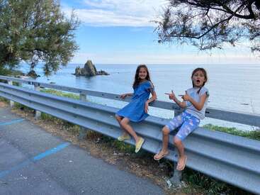 Two young girls sit on a guardrail by the sea, with a serene ocean view and a blue sky with clouds in the background.