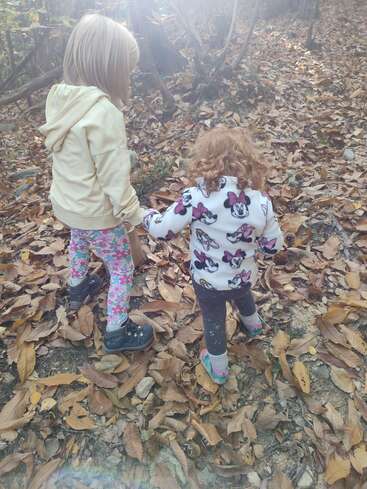 Zwei kleine Kinder, eins blond und eins mit lockigem Haar, laufen Hand in Hand durch einen Wald voller Herbstblätter, angezogen in bunten, kuscheligen Klamotten.