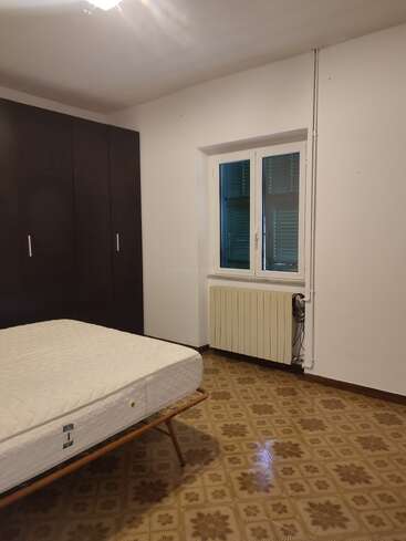 Ein schlichtes Schlafzimmer mit einer Einzelmatratze auf einem Metallgestell, einem gemusterten braunen Bodenbelag, einem großen dunklen Kleiderschrank, einem Fenster mit grünen Fensterläden, einem Heizkörper und weißen Wänden.