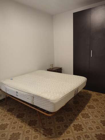 Das Bild zeigt ein schlichtes Schlafzimmer mit einer Einzelmatratze auf einem Metallbettgestell, einem Nachttisch, einem dunklen Kleiderschrank und einem gemusterten Boden. Minimalistische Einrichtung.