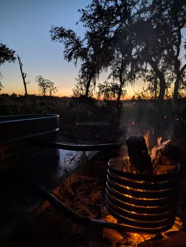 In der Abenddämmerung brennt ein gemütliches Lagerfeuer in einer Metallspirale, umgeben von Bäumen. Der Himmel leuchtet orange und blau, während die Nacht hereinbricht, und strahlt Ruhe aus.