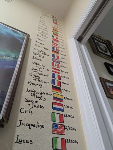 Names and colorful country flags are hand-painted on a wall, each paired with a date, creating a vibrant record of visits or guests over time.
