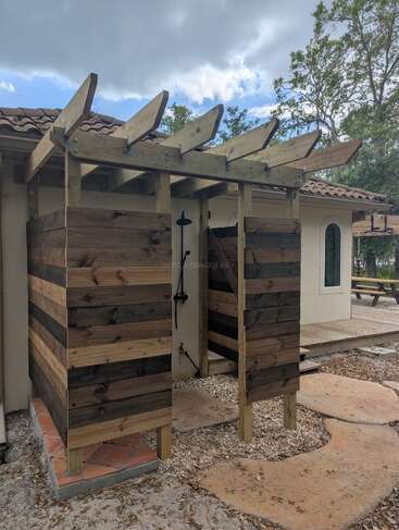 This image shows a rustic outdoor shower made from wooden planks, located next to a house with tile roofing, surrounded by trees and a stone path.