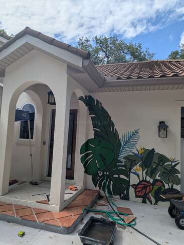 A house entrance with a tiled step, archway, and brown roof. A vibrant tropical plant mural decorates the wall, with tools and hoses scattered around.
