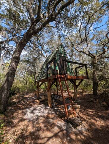 Une charmante cabane dans les arbres en forme de A, construite sur pilotis, se niche au milieu de grands arbres ; dotée d'une échelle en bois et de bardeaux verts, elle est entourée par la nature dans un cadre forestier paisible.