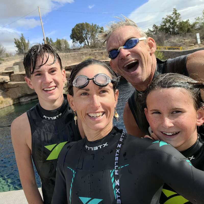 The image shows a family of four wearing wetsuits, posing for a photo near a body of water on a sunny day with a blue sky and white clouds.