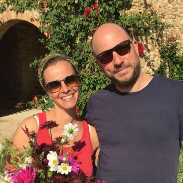 La imagen muestra a un hombre y una mujer juntos, posando para una foto, con la mujer sosteniendo flores delante de un muro de piedra cubierto de vegetación y flores rojas.
