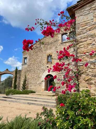 La imagen representa un edificio de piedra con una escalera y un arco, adornado con vibrantes flores rosas y exuberante vegetación, sobre un sereno cielo azul con nubes blancas.