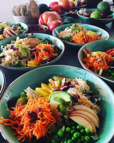 La imagen muestra una mesa repleta de cuencos de ensalada y un surtido de frutas y verduras, en un ambiente culinario vibrante y saludable.