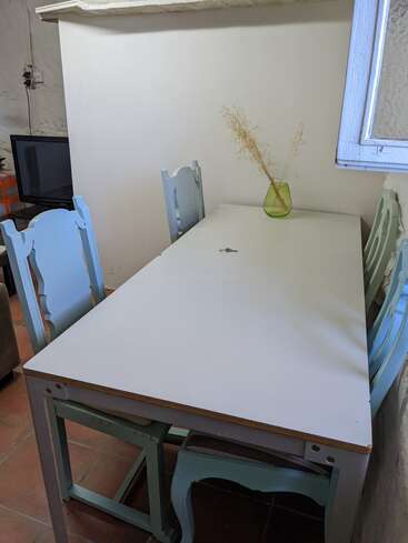 La imagen muestra un pequeño comedor con una mesa blanca y cuatro sillas, un jarrón con flores secas sobre la mesa, una pared blanca y suelo de terracota.