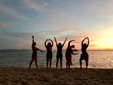 The image depicts five women standing on a beach, forming heart shapes with their arms, silhouetted against a vibrant sunset sky and calm ocean waters.