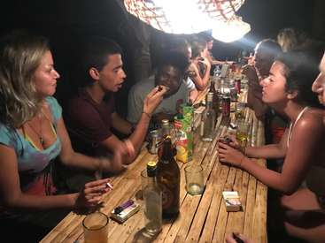 The image depicts a group of people gathered around a long wooden table, enjoying drinks and socializing in a dimly lit setting.
