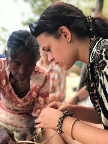 The image shows a woman with dark hair and a black-and-white patterned shirt, assisting an older woman with dark hair and a red-and-white patterned shirt, possibly with a craft project.