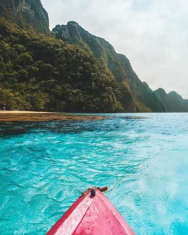 The image depicts a serene scene of a red boat navigating through crystal-clear turquoise waters, with lush green mountains and a sandy beach visible in the background.