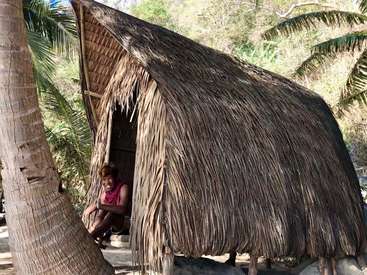 The image depicts a woman sitting in the doorway of a hut with a thatched roof, surrounded by palm trees and lush greenery.