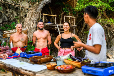 Four people, two shirtless men and a smiling woman, participate in an outdoor cooking class. Fresh vegetables and stoves sit on a rustic wooden table, jungle background.