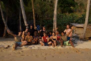 A diverse group of people sits joyfully on a sandy beach, surrounded by palm trees and greenery, smiling and waving, with a hut in the background.