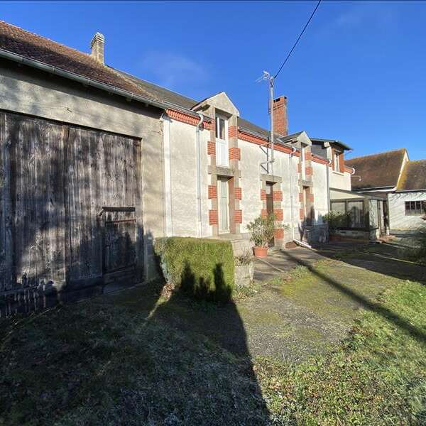 Una casa de campo rústica con paredes blancas y detalles de ladrillo rojo se alza bajo un cielo azul despejado. Hay una puerta de madera de granero y un patio cubierto de hierba.