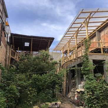 The image shows a construction site with partially built bamboo and brick structures, surrounded by overgrown greenery, under a blue sky with scattered clouds.