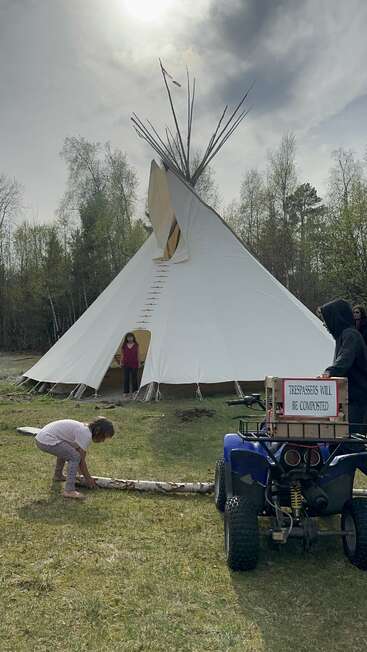 En un claro cubierto de hierba se alza un gran tipi blanco. Hay tres personas: un niño con un tronco y un quad con un cartel que dice: «Los intrusos serán convertidos en abono».
